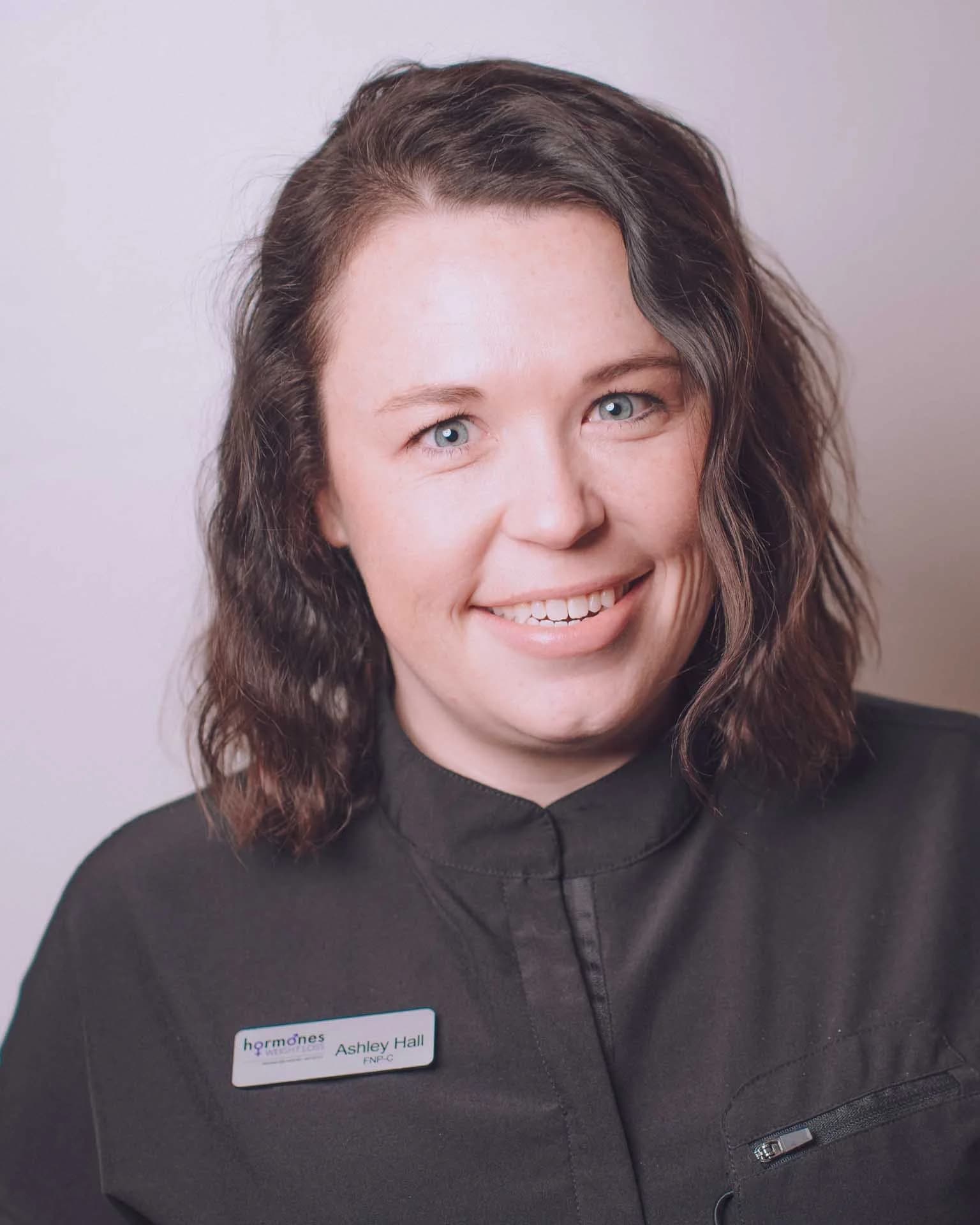 A woman with shoulder-length wavy brown hair, wearing a black uniform with a name tag that reads 'Ashley Hall, FNP-C,' smiling against a plain background.