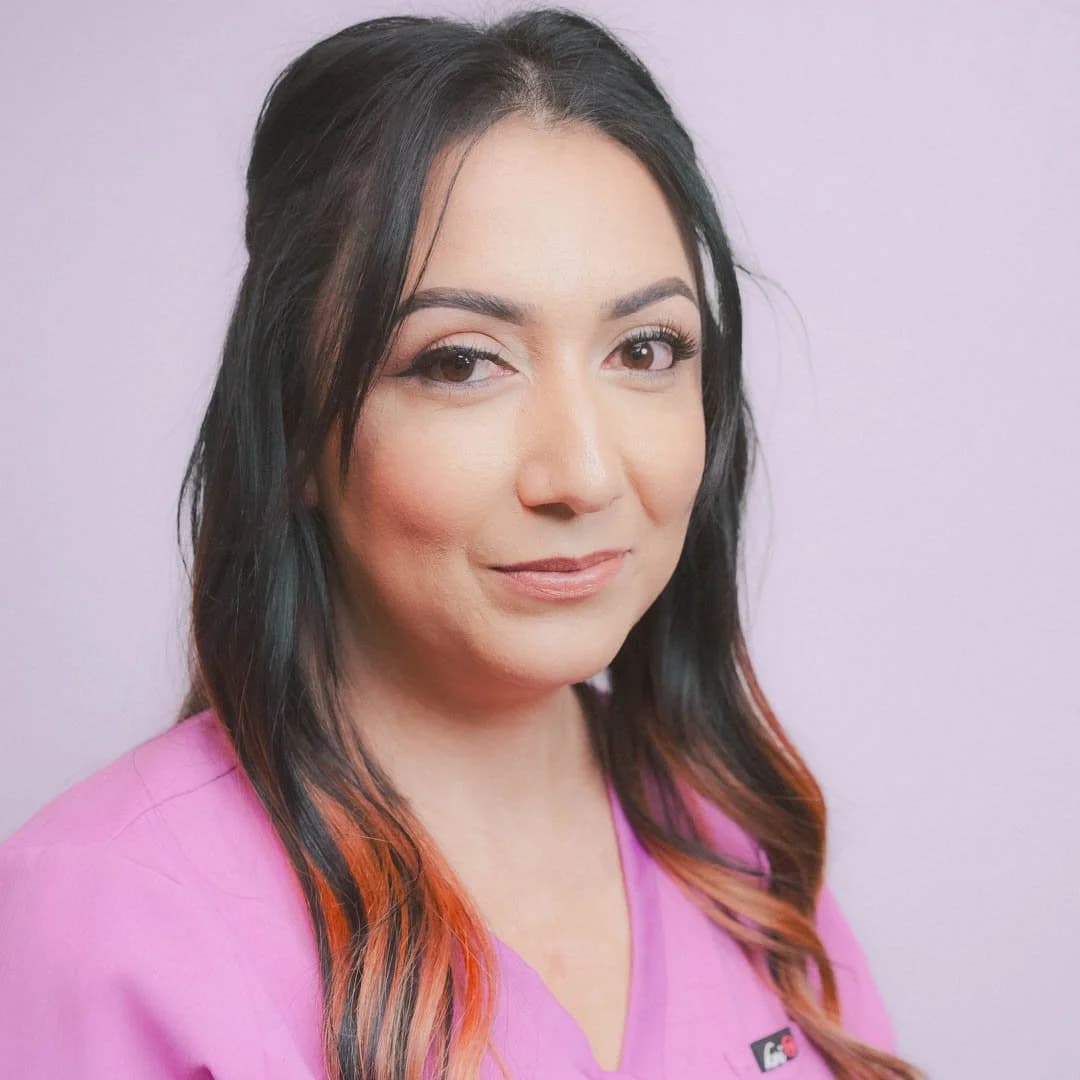Portrait of a woman with dark hair, some reddish ends, wearing a pink top, standing against a light purple background.