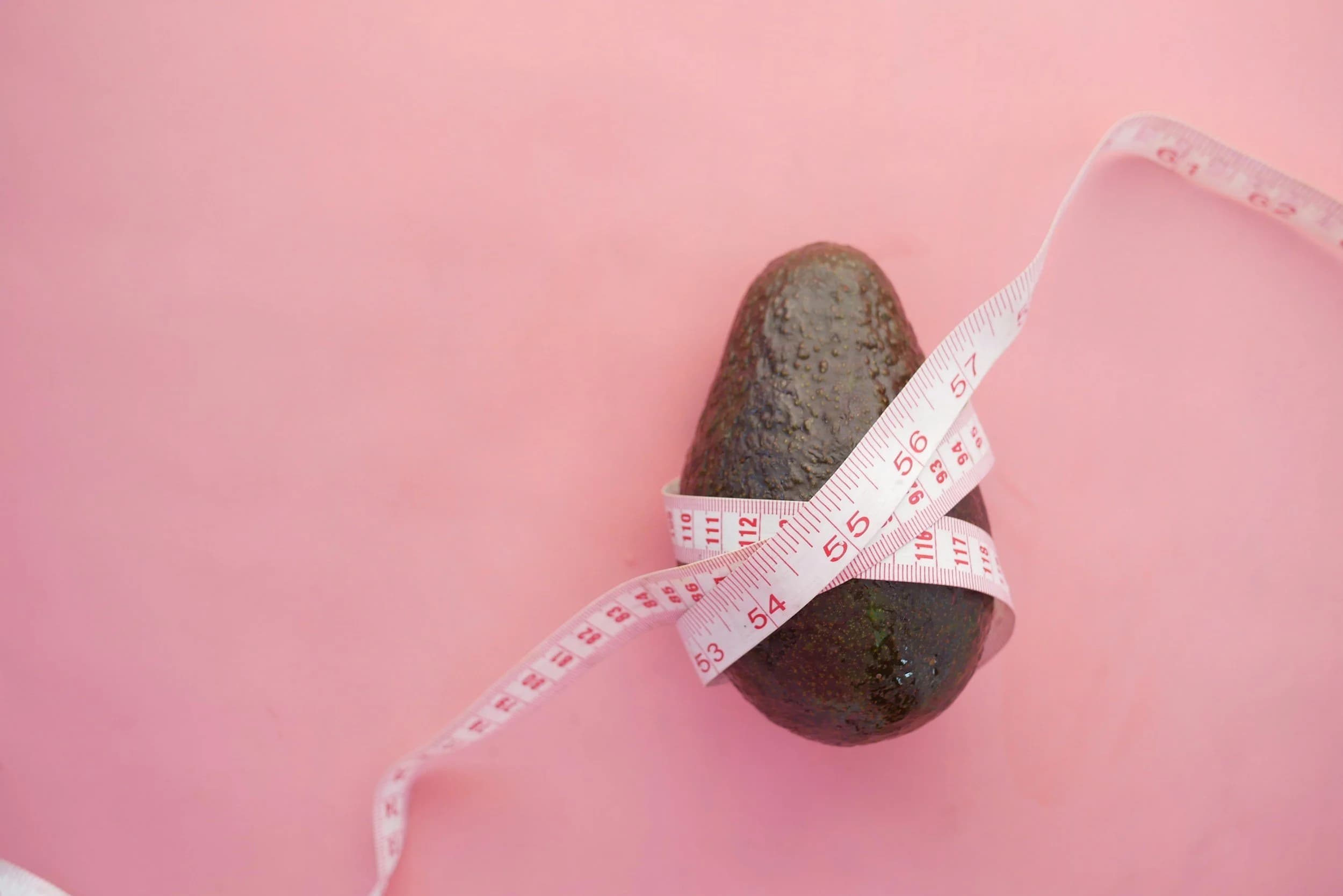 An avocado is wrapped with a pink measuring tape on a pink background.