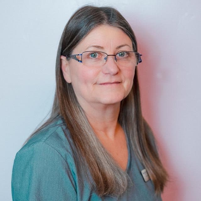 A woman with long brown hair, glasses, wearing a teal top, standing against a white and light pink background.
