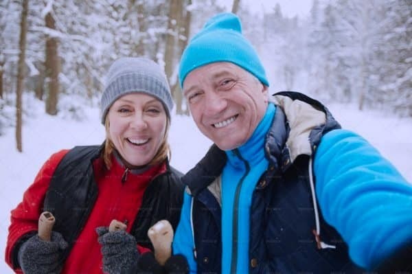 A smiling man and woman in winter clothing taking a selfie in a snowy forest.